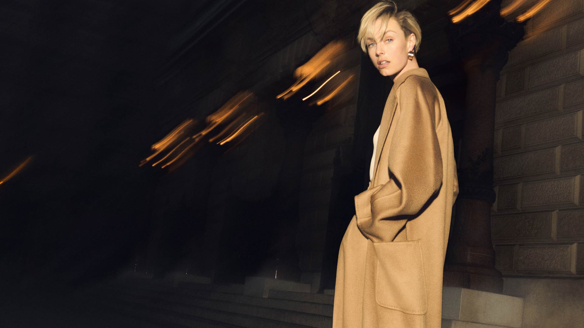 Modelka w obszernym, kultowym płaszczu Ludmilla Max Mara w kolorze wielbłądzim, w wieczorowej scenerii.